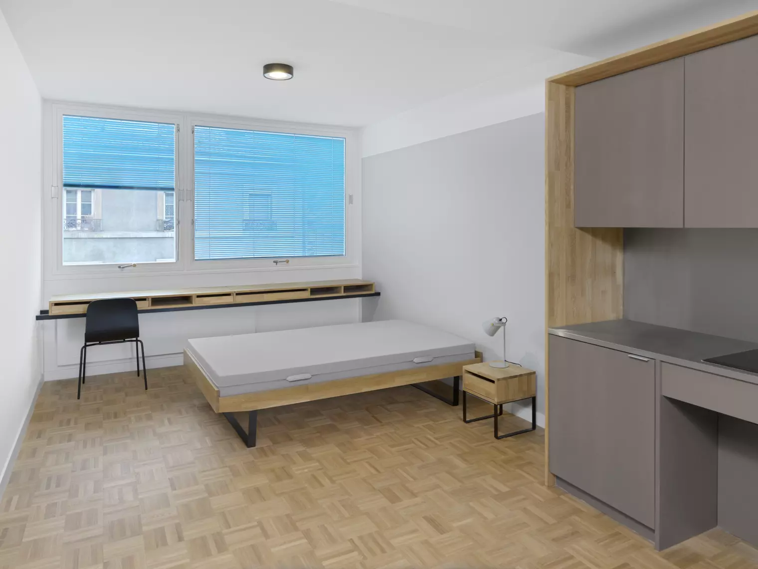 Un studio avec une console en bois et une chaise, une grande baie vitrée, un lit et une table de chevet et une kitchenette. Crédits photo : Annik Wetter
